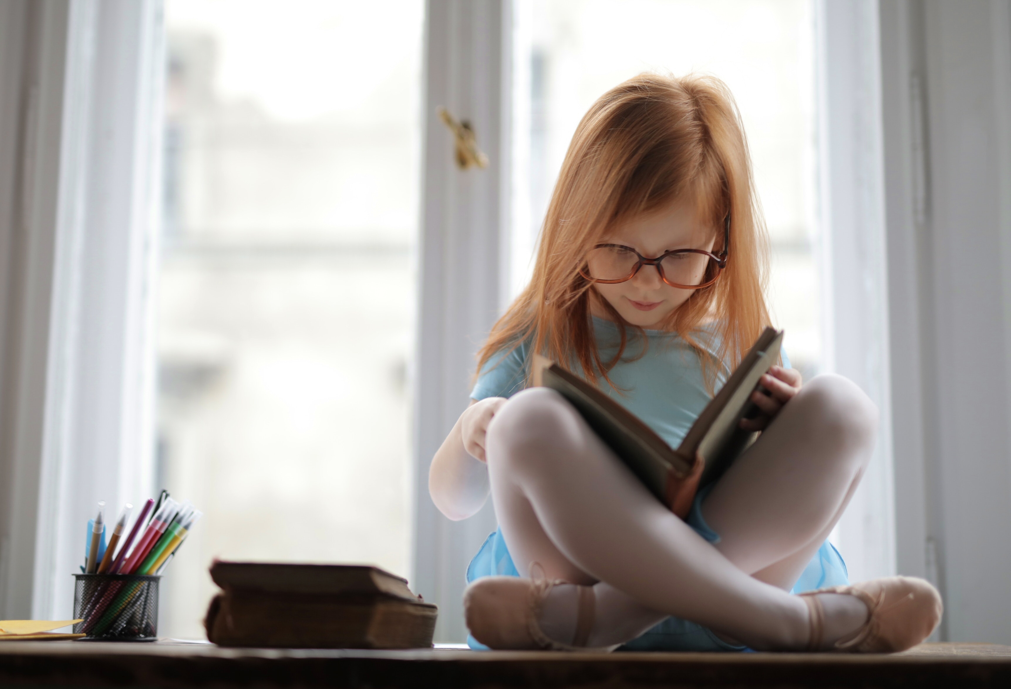 Na foto, uma menina ruiva, de cabelos lisos, está sentada em cima de uma mesa, lendo um livro, com outros livros do lado e muitos lápis em um potinho. A foto foi tirada em frentre a uma janela.