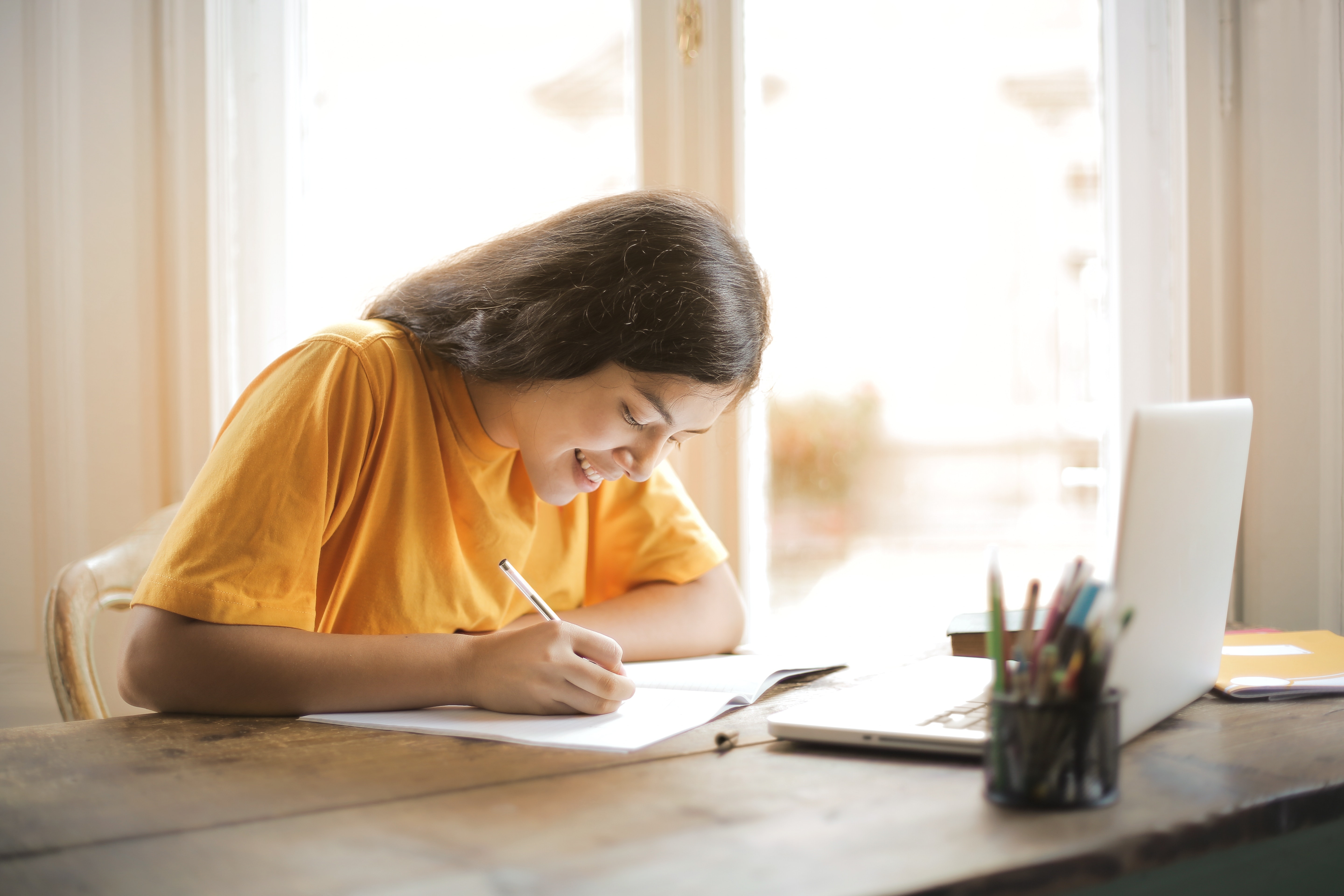 Imagem mostra uma menina escrevendo em um folha e um notebook a frente
