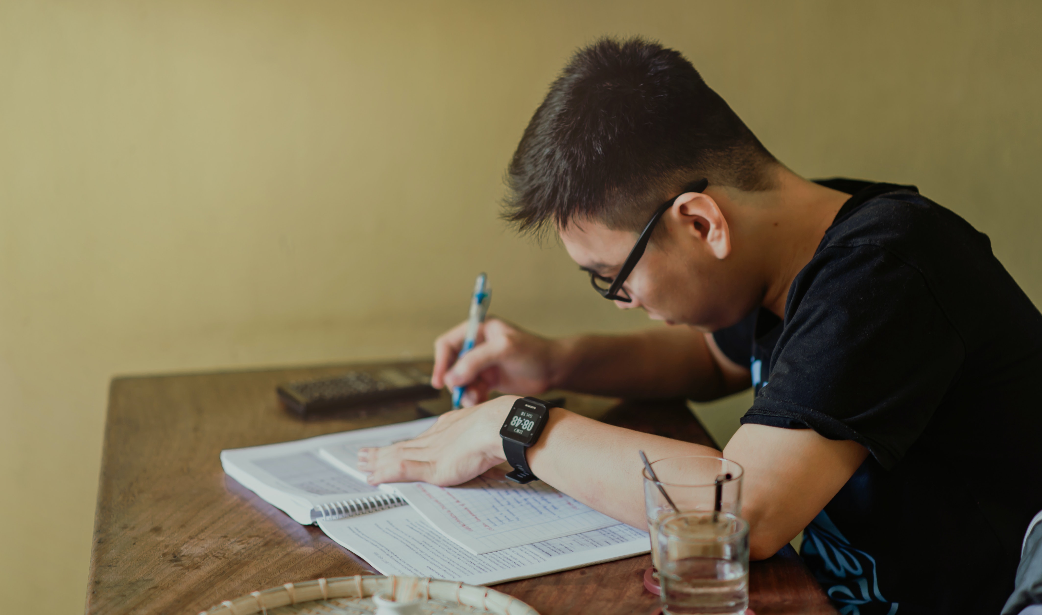 Imagem mostra um menino adolescente de lado,  com uma caneta nas mãos escrevendo em uma apostila. 