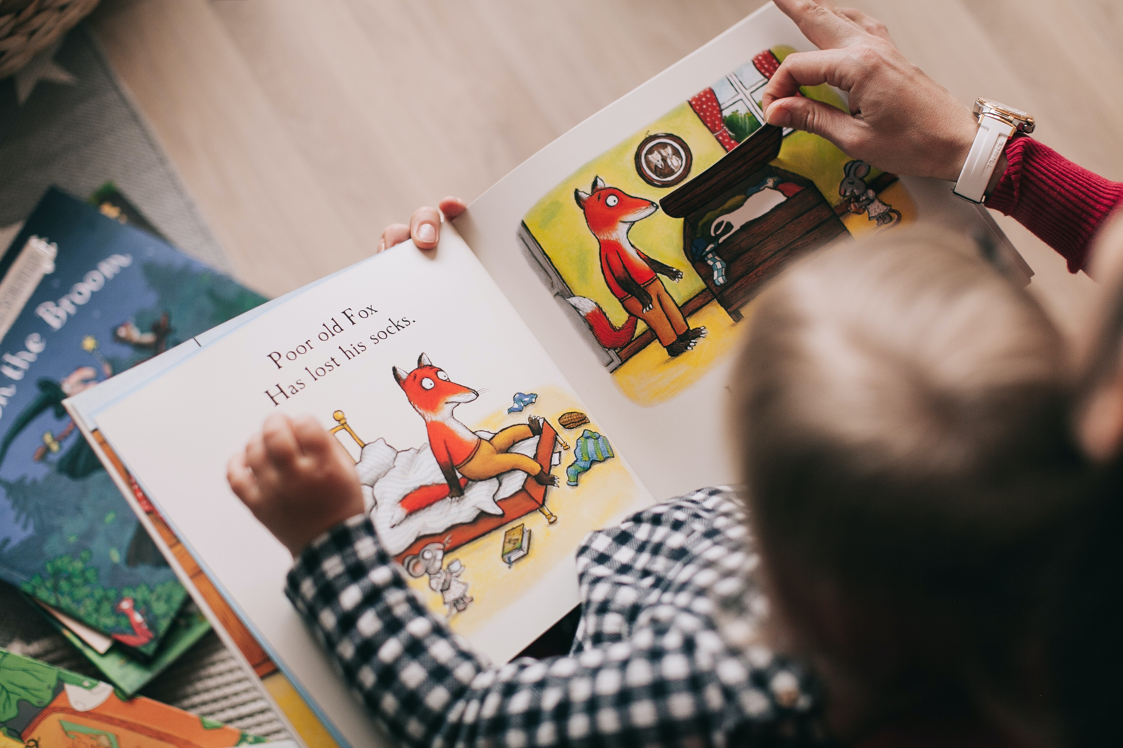 A foto foi tirada de cima e mostra a cabeça de uma criança e suas mãos em cima de um livro, com ilustrações de uma raposa. A criança está acompanhando por um adulto, porque conseguimos ver a mão de uma mulher adulta, ajudando a criança na leitura.