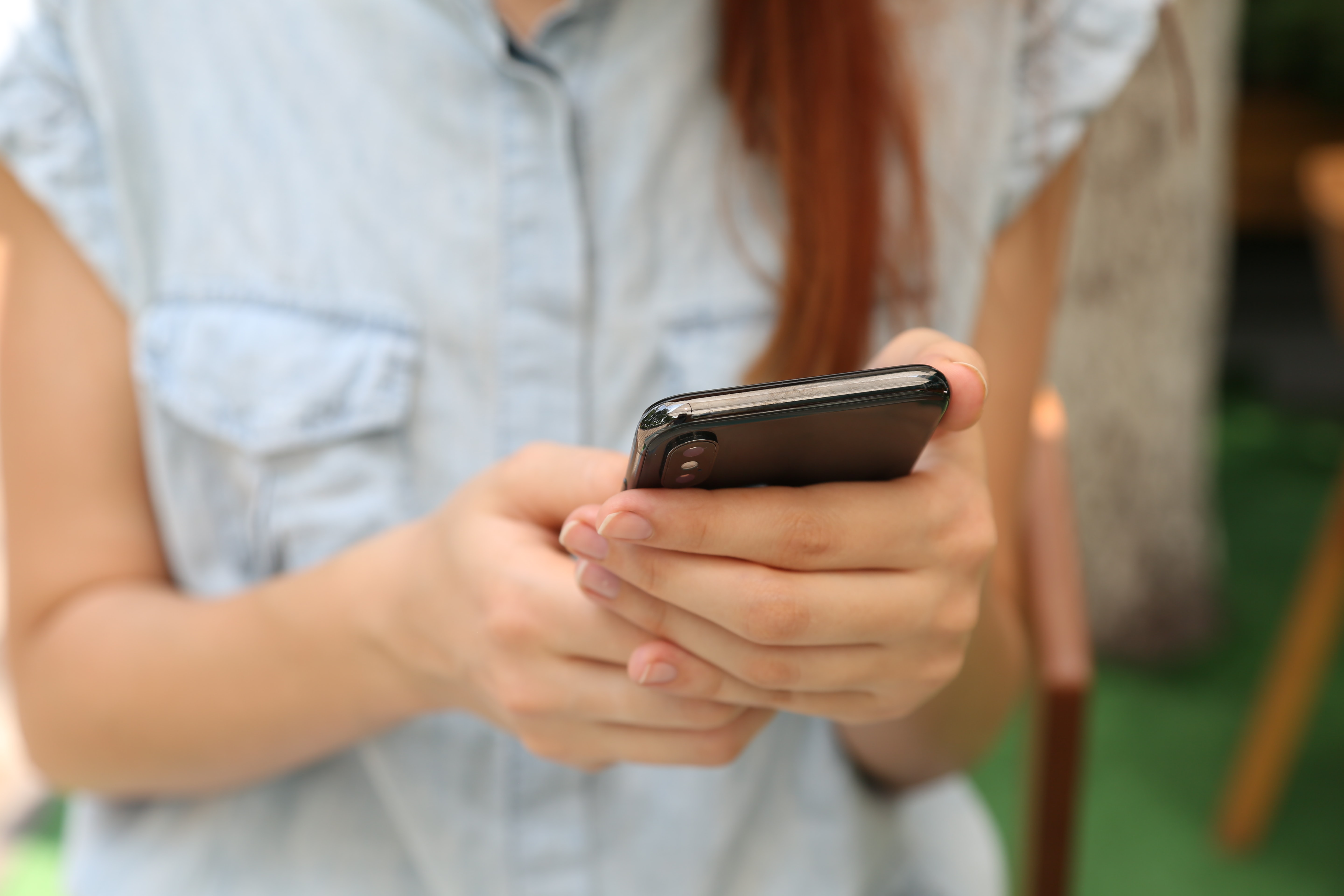 A foto mostra uma mulher com cabelo comprido mexendo no celular. Seu rosto não aparece, apenas seus braços e as mãos segurando o celular. 