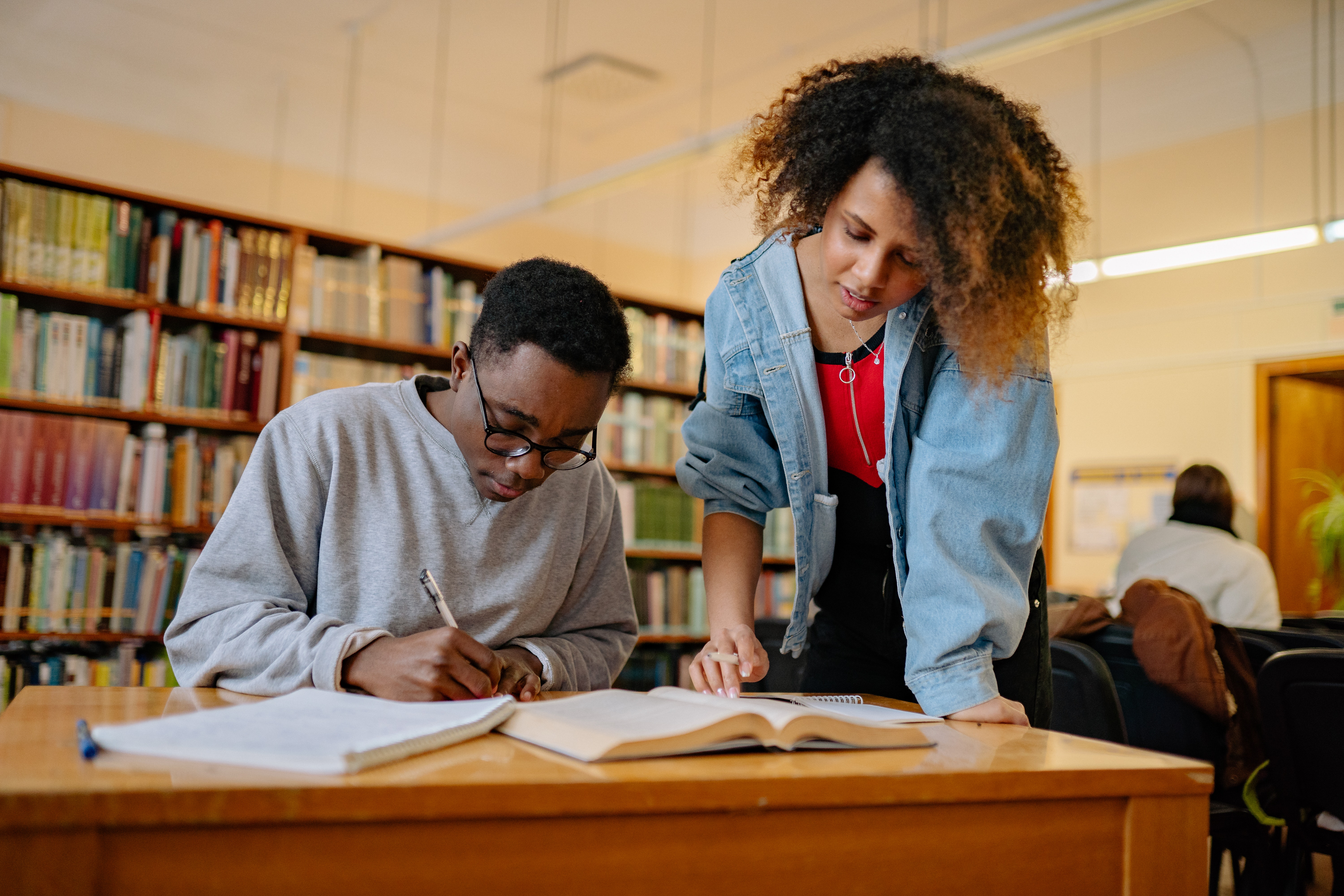 Um homem negro está sentado escrevendo e uma mulher negra está em pé olhando os materiais ao seu lado. Eles estão em uma biblioteca. 