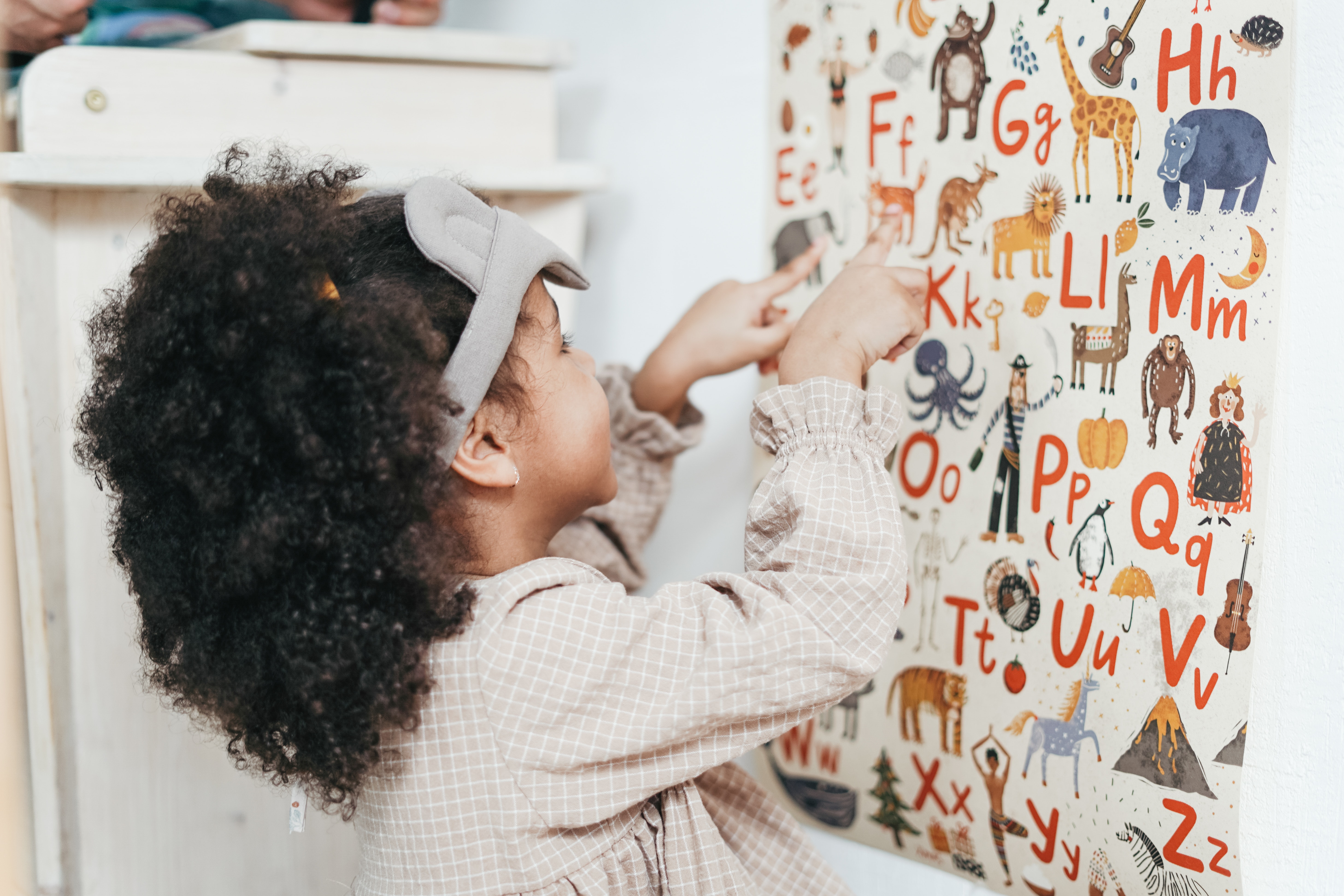Uma menina de aproximadamente quatro anos está em frente a um painel cheio de letras do alfabeto. Todas as letras têm ao lado um animal, como girafa, elefante. 