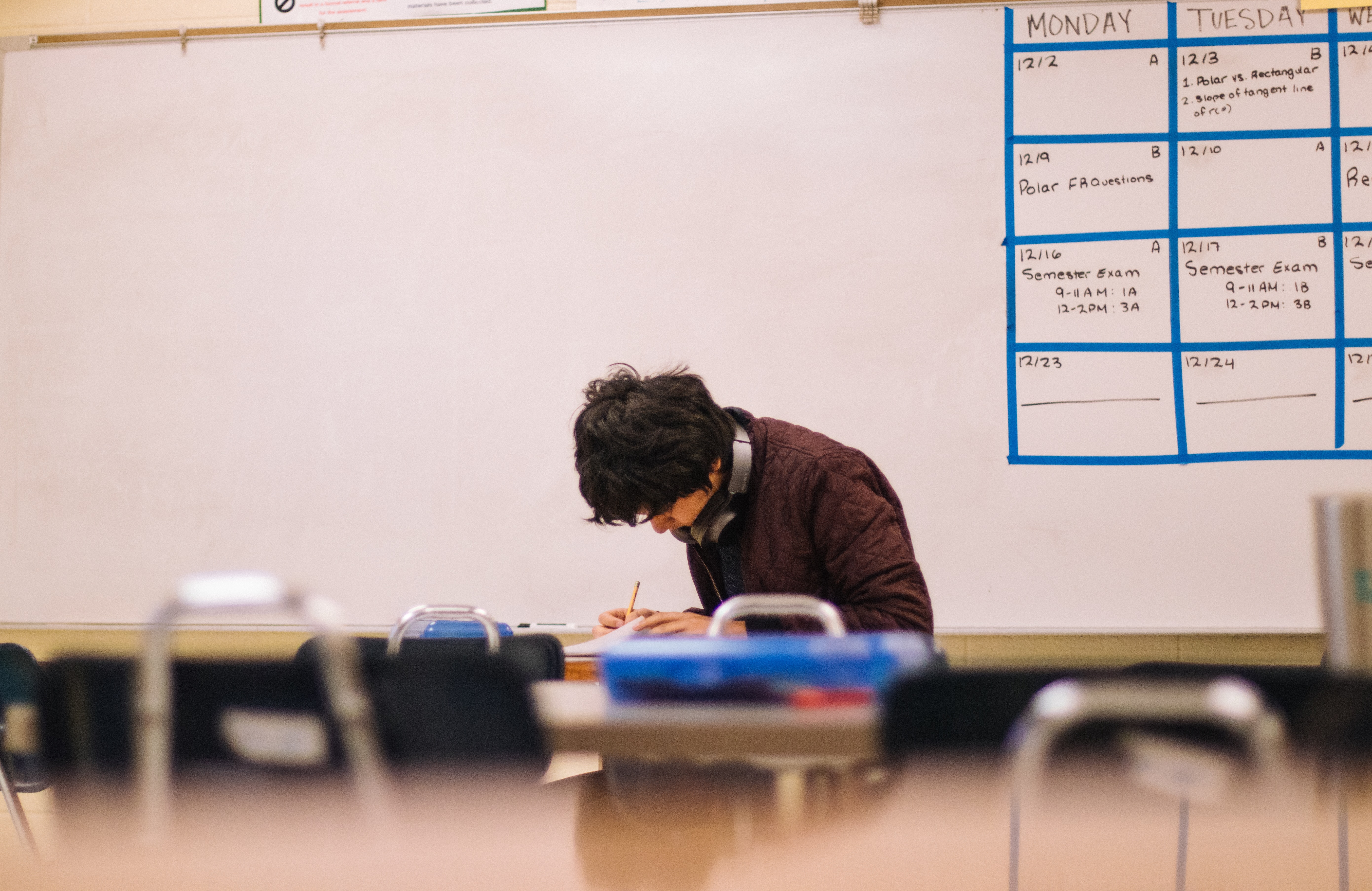 A foto, tirada de longe, mostra menino fazendo uma prova em uma sala de aula. Atrás podemos ver uma lousa. 