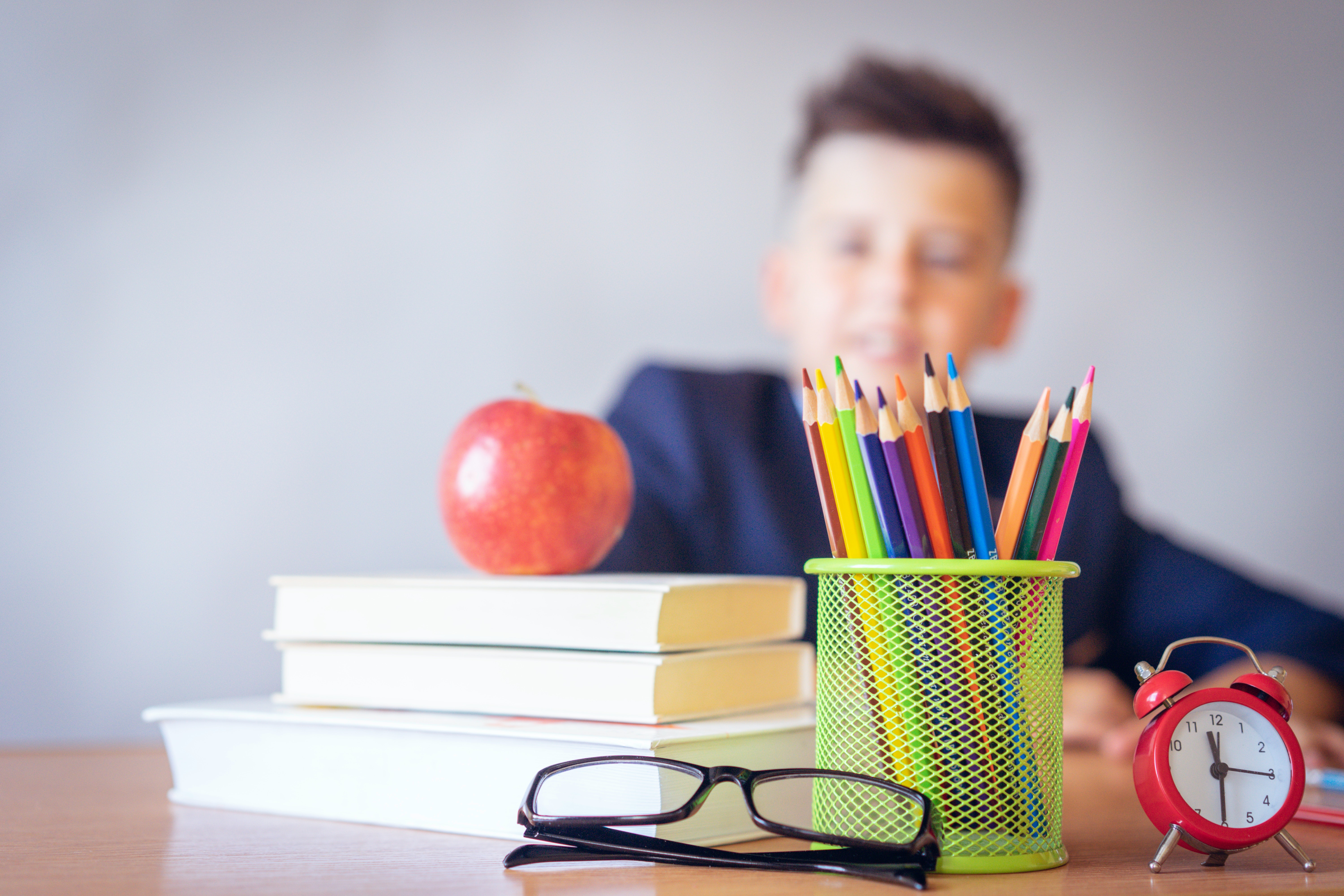 Professor sentado em sua mesa com livros, canetas, relógio e uma maçã.