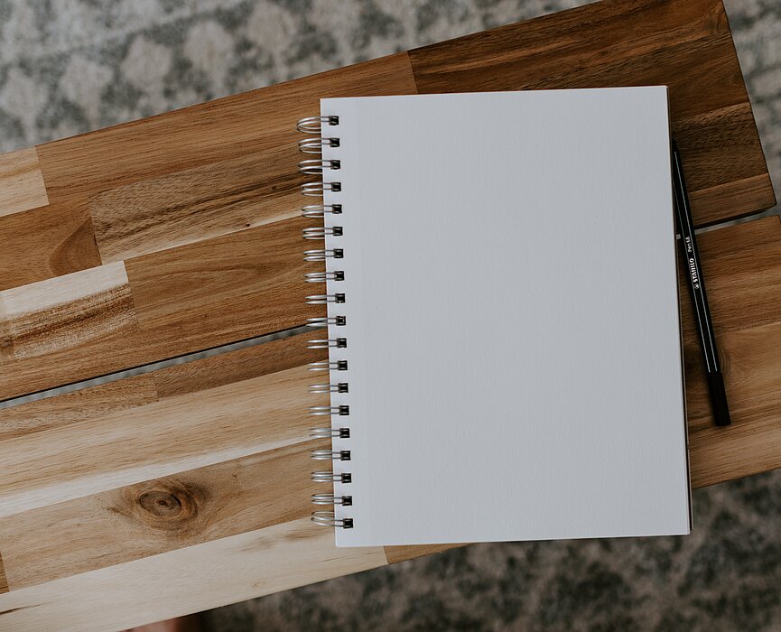 A foto mostra um caderno em cima de uma mesinha de madeira. Ao lado tem uma caneta. 