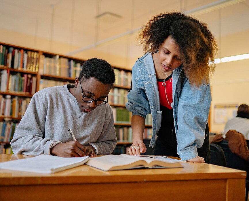 Um homem negro está sentado escrevendo e uma mulher negra está em pé olhando os materiais ao seu lado. Eles estão em uma biblioteca. 