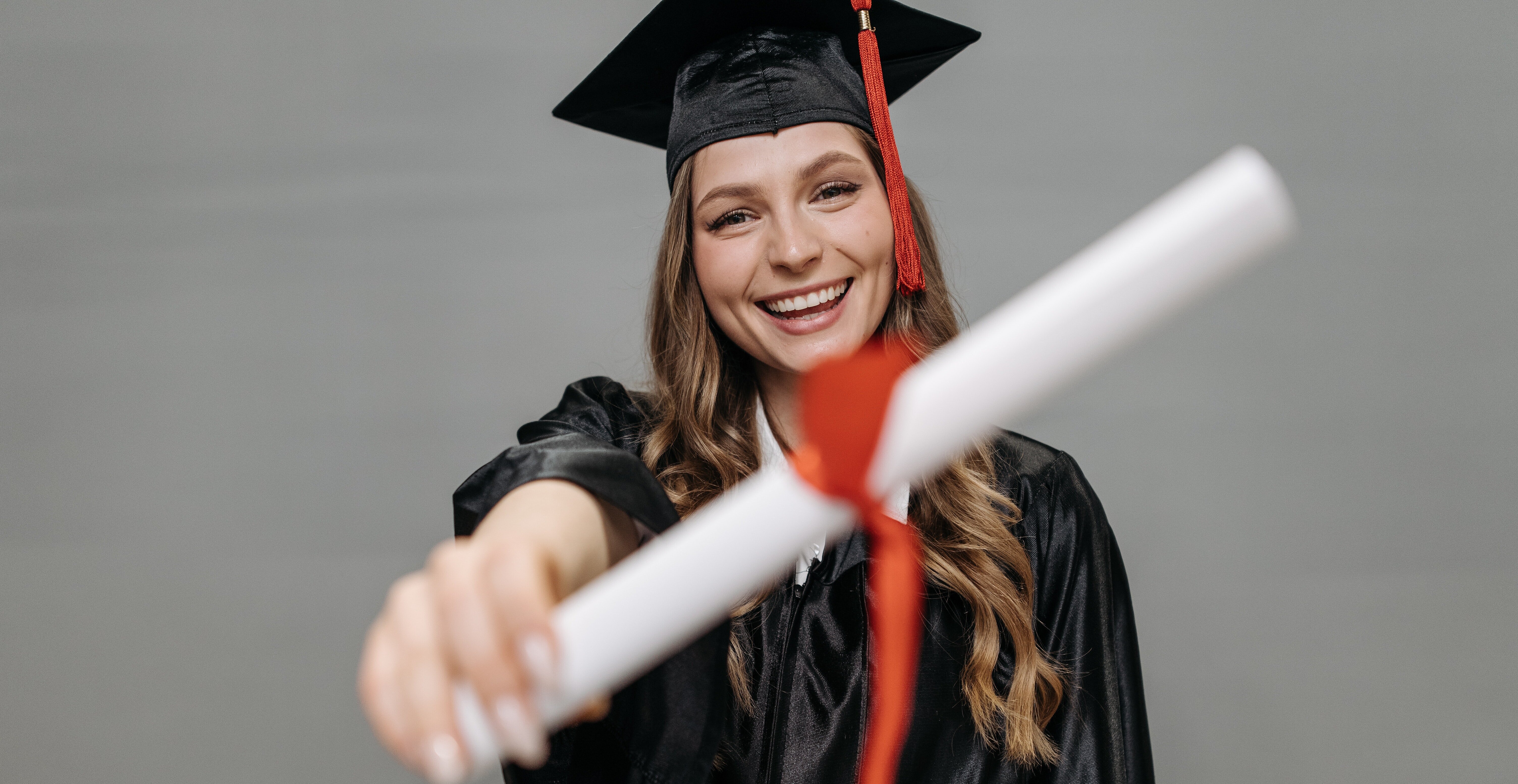 Na foto, uma menina mostra seu diploma para a câmera. Ela está de beca, com capelo e está sorrindo.