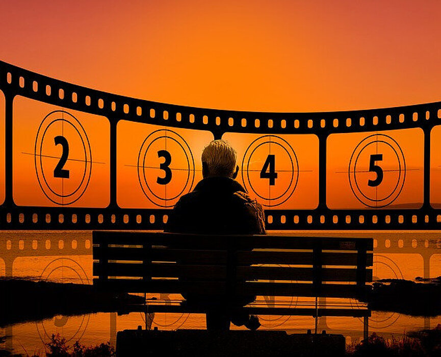 Idoso sentado em um banco de parque, em frente a um lago, com horizonte com vista de rolo de filmes numerado. Foto preta e laranja.