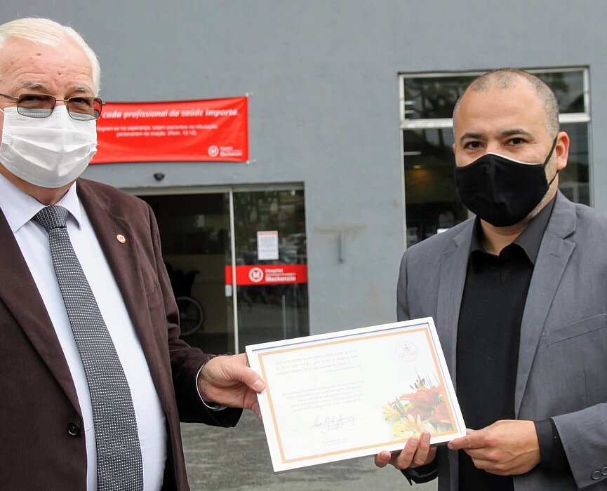 Dois homens com máscaras e ternos seguram um quadro de reconhecimento em frente ao hospital