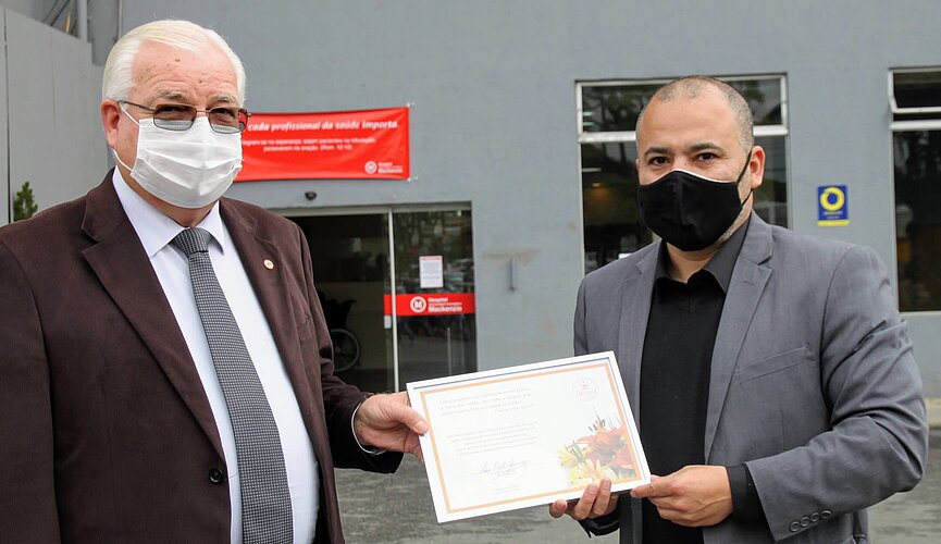 Dois homens com máscaras e ternos seguram um quadro de reconhecimento em frente ao hospital