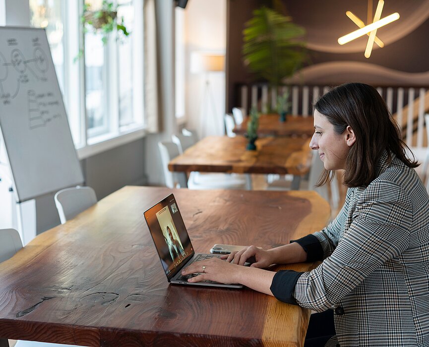 Uma mulher está sentada em uma mesa, em frente a um notebook. O espaço parece ser um café, mas ela está sozinha. Do lado esquerdo da foto podemos ver um quadro, com algumas coisas escritas. 