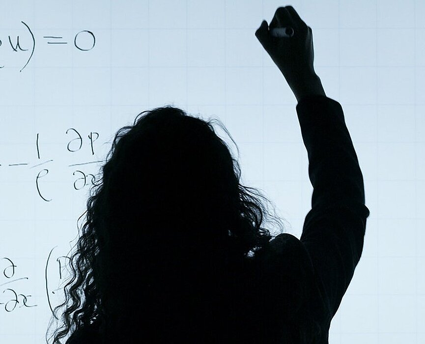 Mulher de cabelos longos, de costas, escreve equações em uma lousa branca