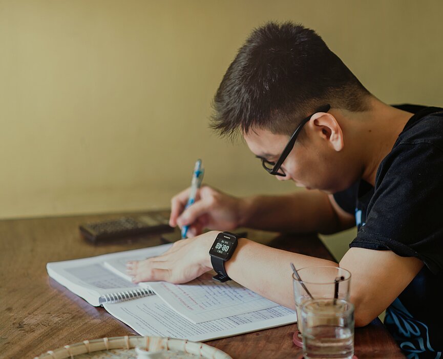 Imagem mostra um menino adolescente de lado,  com uma caneta nas mãos escrevendo em uma apostila. 
