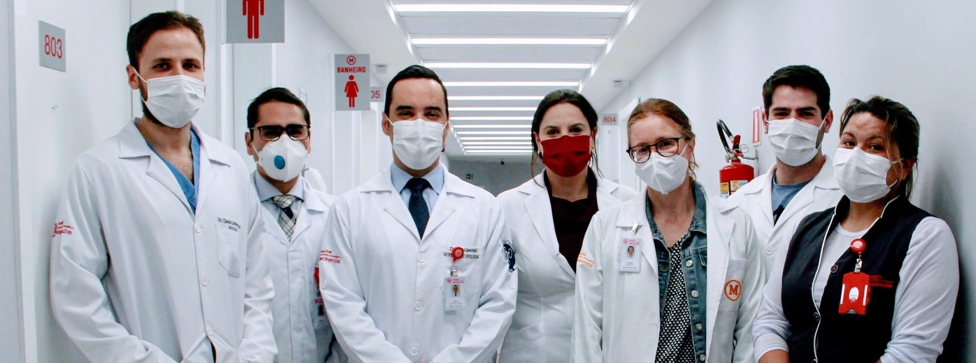 Na imagem, sete pessoas entre homens e mulheres, posam para foto num corredor claro de hospital. As pessoas vestem jalecos e uniformes com símbolo do Mackenzie e estão usando máscaras