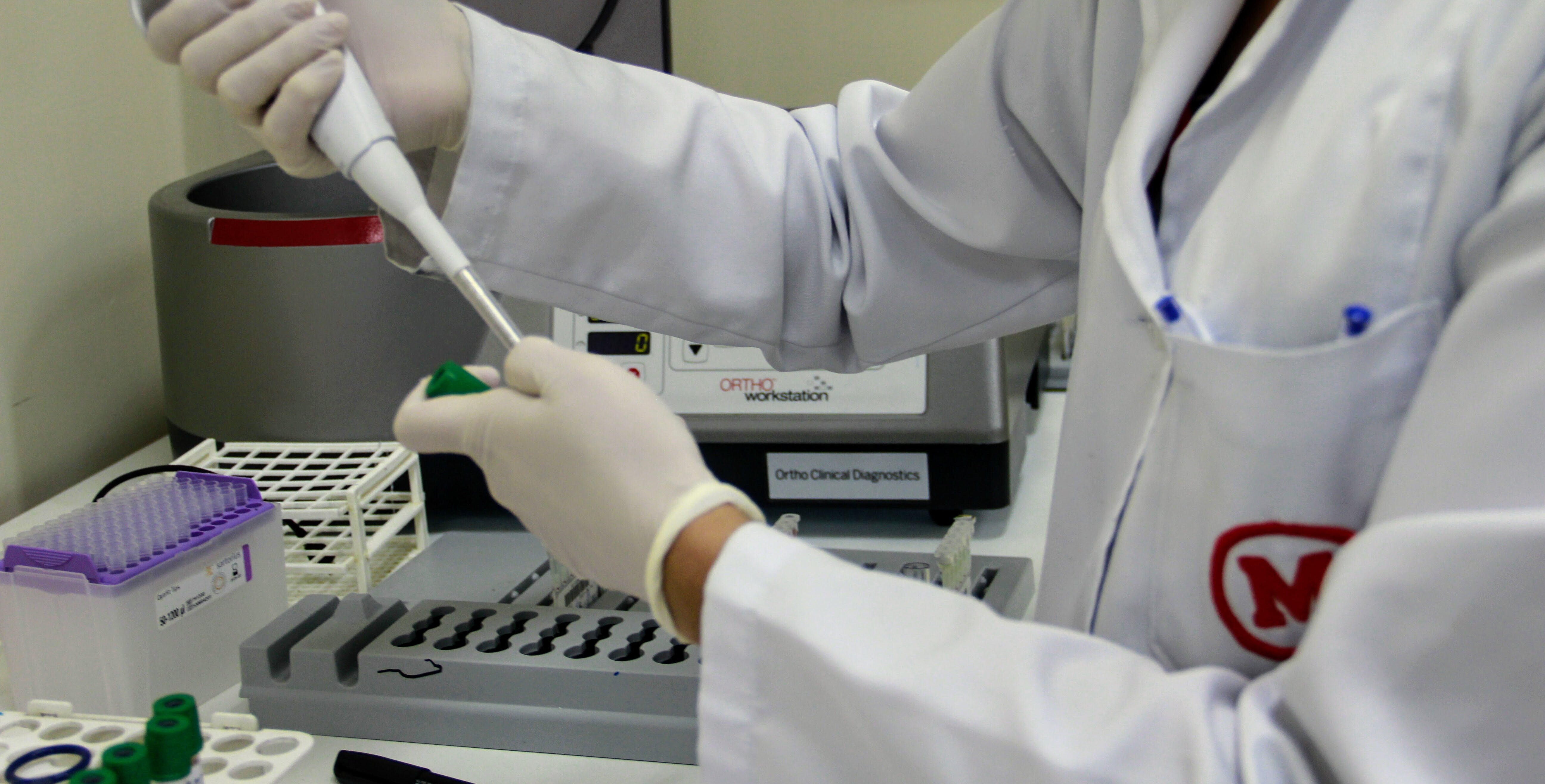 profissional de laboratório com jaleco branco com símbolo do Mackenzie em vermelho, manipulando uma pipeta de laboratório